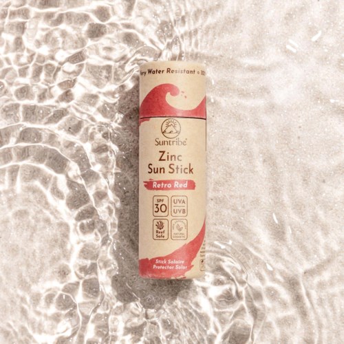 Słoik Suntribe Zinc Sun Stick SPF 30 leży na piasku, częściowo w wodzie, wskazując na użycie ochrony przed słońcem.