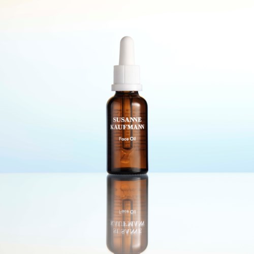 En brunt flaska med vit pipettlockmärkning "Susanne Kaufmann Face Oil." Reflektion syns nedan.