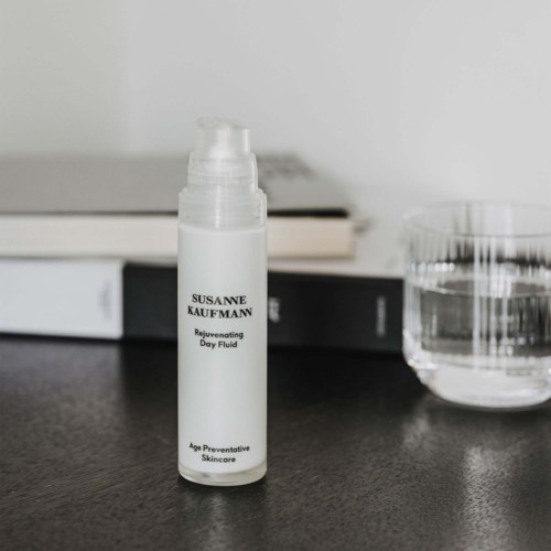En vit flaska märkt 'Rejuvenating Day Fluid' och 'Age Preventive Skincare' på en svart yta med böcker och ett glas vatten.