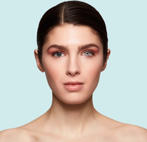 Eine Frau mit heller Haut, braunen Haaren und natürlichem Make-up vor einem hellblauen Hintergrund.