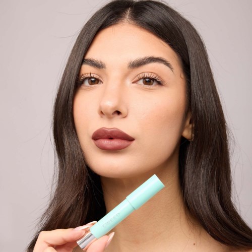 A woman with long dark hair and full lips holds a light blue tube, possibly a beauty product, in front of her face.