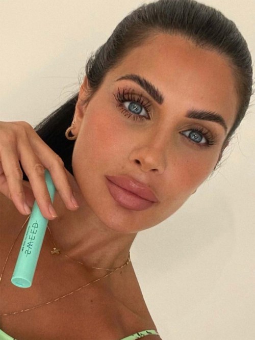 A woman with bright blue eyes, long dark hair, and full lips holds a blue tube. She wears makeup and earrings.