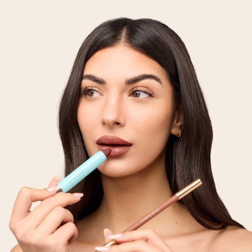 A woman with long dark hair applies lipstick and holds a tube of makeup.