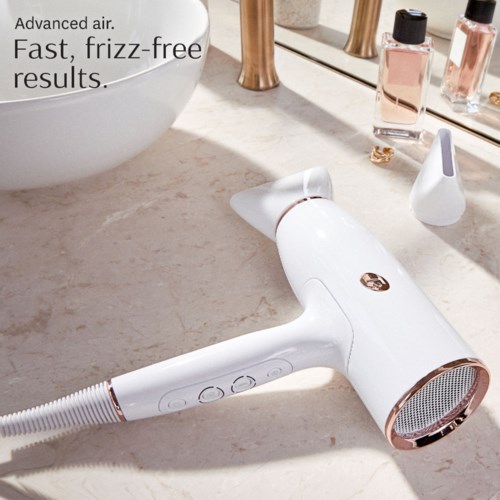 A white hairdryer with rose gold accents lies on a beige countertop near bottles of hair products, with a mirror in the background.