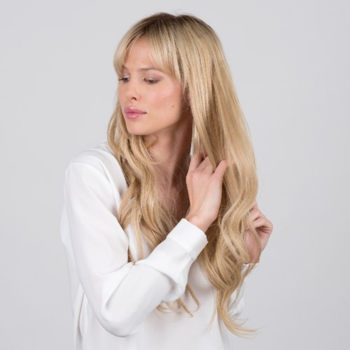 A woman with long blonde hair touches her hair, wearing a white top, looks down, neutral expression.