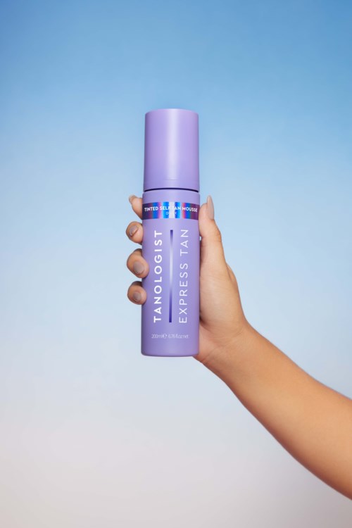Dłoń trzyma fioletową butelkę spray z napisem "TANOLOGIST EXPRESS TAN" na niebieskim tle.