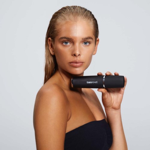 Een vrouw met blond haar houdt een zwart cilindrisch voorwerp bij haar schouder, kijkt recht vooruit, met een neutrale achtergrond.