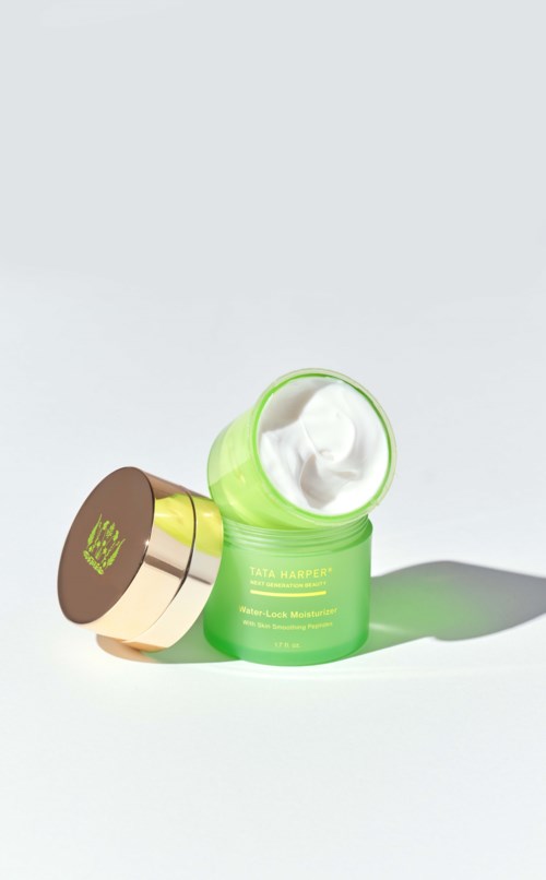 A green jar with a gold lid, open to show white cream inside, labeled Tata Harper. Light background.