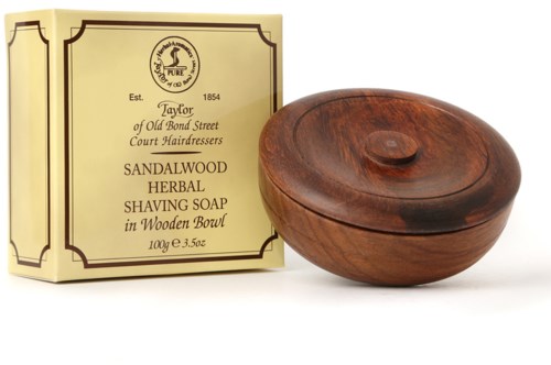 A wooden bowl with a lid next to a cream-colored package of herbal shaving soap. The package has text and a logo.
