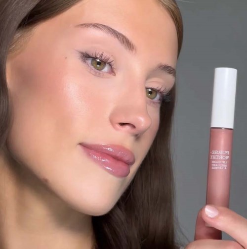 Een jonge vrouw met glanzende lippen en een egale huid houdt een lippenstift vast, glimlacht vriendelijk.