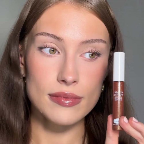 Een vrouw met lang bruin haar toont een lipglossfles, kijkt lichtjes zijwaarts.