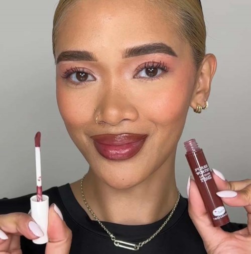 Een vrouw met blond haar, make-up, houdt een lipgloss en een lippenstift vast, lacht naar de camera.