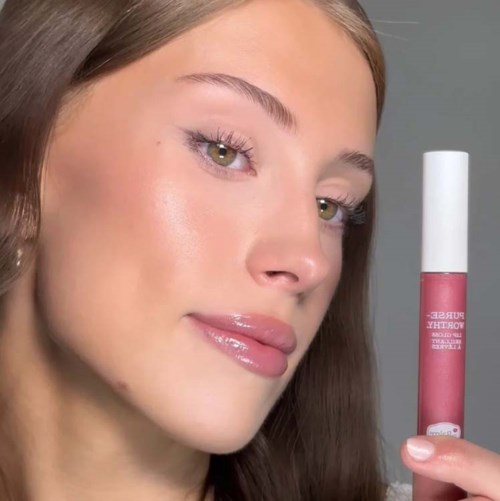 Een vrouw met glanzende lippen en een heldere huid houdt een roze lipgloss. Ze heeft lang bruin haar en lichte ogen.