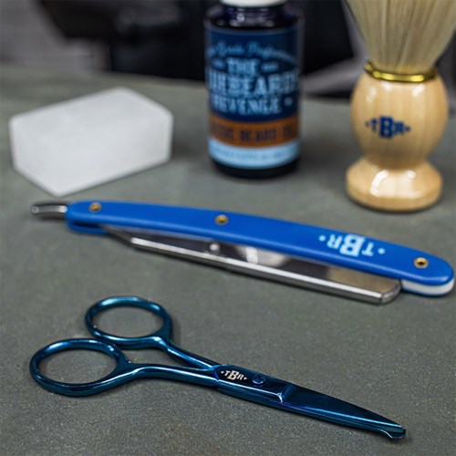 A blue pair of small scissors and a blue-handled nail clipper on a gray surface. A white soap, a blue bottle, and a shaving brush in the background.