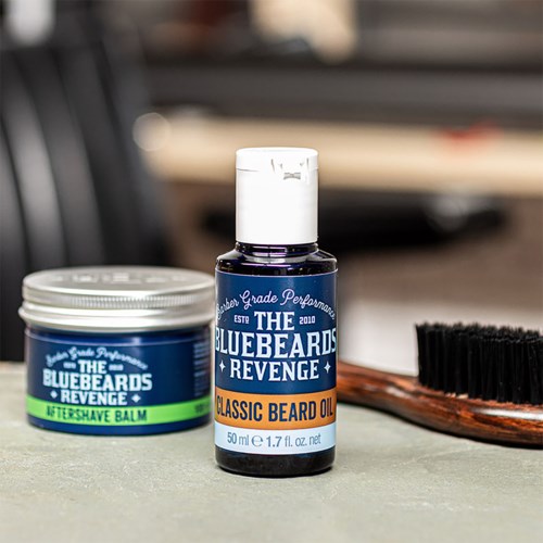 A small bottle of beard oil, a container of beard balm, and a brush are on a gray surface.