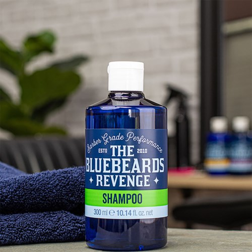 Blue bottle labeled 'Shampoo' next to a folded towel, outdoor background with some bottles. The label mentions performance and revenge.