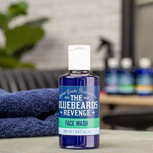 A bottle labeled 'The Bluebeards Revenge Face Wash' on a desk, with a towel and barber supplies in the background.