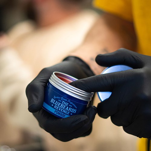 A person wearing black gloves holds a small round blue container of pomade, with part of the lid visible.