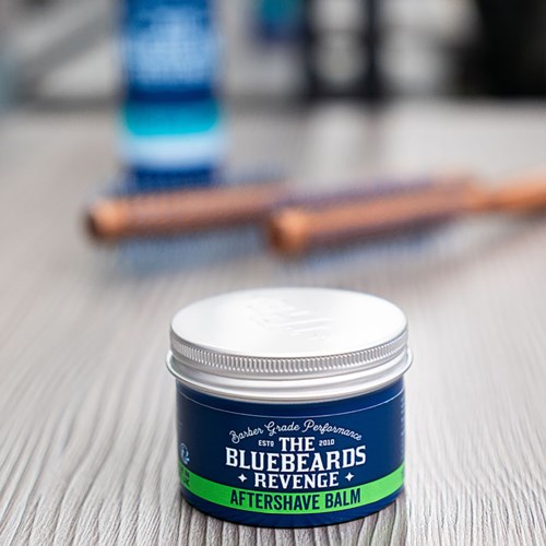 A blue jar labeled 'Revenge Aftershave Balm' sits on a table with blurred grooming tools in the background.