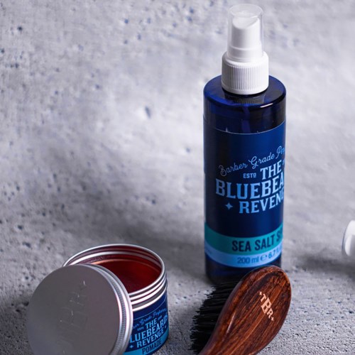 Eine blaue Sprühflasche, ein kleiner offener Behälter, eine Holzborste und eine weiße Wand.