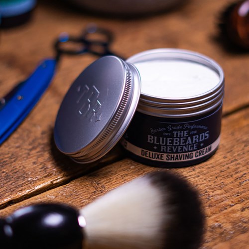 A jar of Bluebeards Revenge shaving cream on a wooden surface with grooming tools nearby.