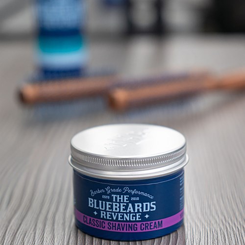 A jar of Bluebeard's Revenge Classic Shaving Cream on a wooden surface with blurred background objects.