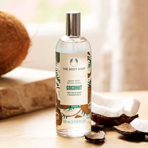 Eine transparente Sprühflasche mit Kokos-Body-Mist auf einer Holzoberfläche, mit Kokosnussschalen und weichem natürlichem Licht.