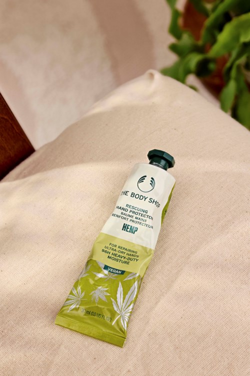 Een tube hennep handhulpcreme op beige stof met groene planten op de achtergrond.