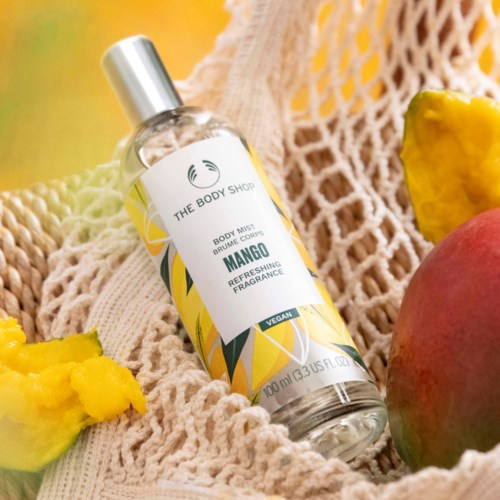Ein Mango-Duft Körperspray in einer transparenten Flasche auf einem beigefarbenen Tuch mit Mango- und Ananasstücken daneben.