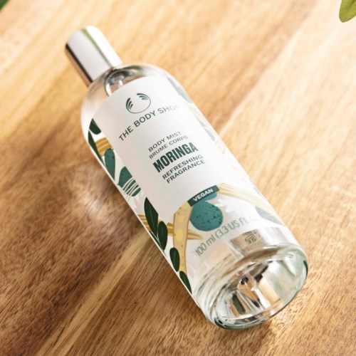 Eine klare Sprühflasche mit Deckel, beschriftet mit 'Moringa Refreshing Fragrance', liegt auf einer Holztisch.