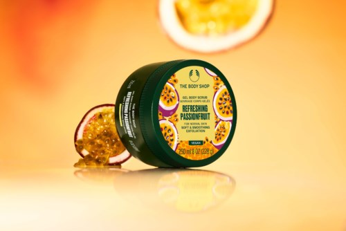 Ein grüner Behälter von The Body Shop Passionsfrucht Peeling mit Passionsfruchthälften daneben, auf gelbem Hintergrund.