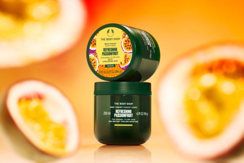 Zwei kleine Behälter mit Körpercreme mit Passionsfruchtduft, umgeben von Passionsfrucht-Hälften, auf orangefarbenem Hintergrund.