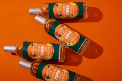 Four orange bottles of pumpkin body oil with silver caps are arranged on an orange background, casting shadows.