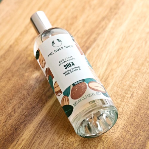 Eine transparente Wassersprühflasche mit der Aufschrift 'Shea' und bunten abstrakten Kunst auf einer Holzoberfläche.