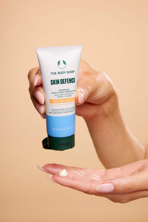 A hand with light skin holding a tube of The Body Shop Skin Defence moisturizer on a beige background.