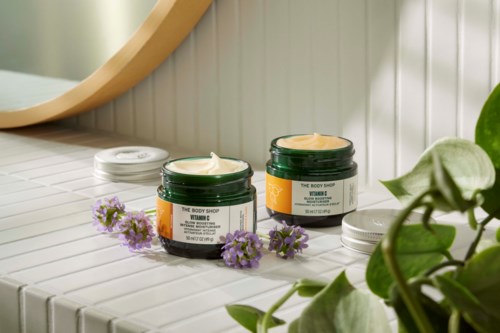Zwei grüne Gläser mit Creme, umgeben von violetten Blumen und grünen Blättern, auf weißer Fliesenfläche.