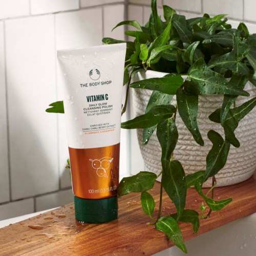 En vit tub från The Body Shop Vitamin M Bright Glow-rengöring bredvid en grön växt på en träytan.