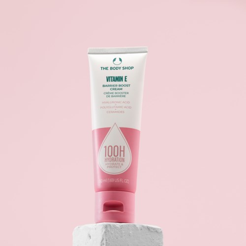 Das Bild zeigt eine rosa-weiße Tube von The Body Shop Vitamin E Creme auf einem weißen Ständer vor rosa Hintergrund.