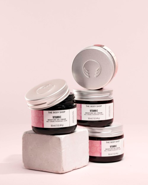 Three jars of body cream labeled Vitamine are stacked on a white block with a pink background.
