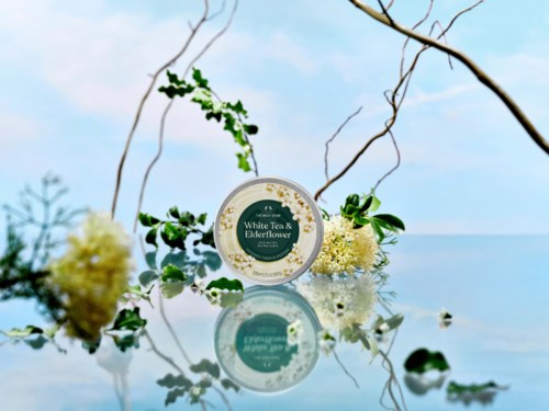 Ein runder Behälter mit der Aufschrift White Tea Elderflower, umgeben von Blumen und grünen Blättern, mit Himmel- und Wasserreflex im Hintergrund.