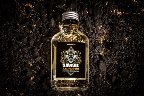 A small perfume bottle labeled "SAVAGE" with a roaring lion emblem, placed on black volcanic rock. Gold accents visible.