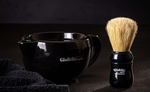 A black shaving bowl and brush with a black towel, all with the Goodfellas smile logo, on a dark surface.