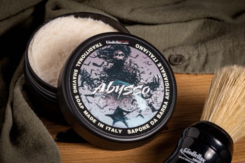 En svart rund beholder med barberkrem med en skalle og trelogg, ved siden av en barberbørste på treplate.