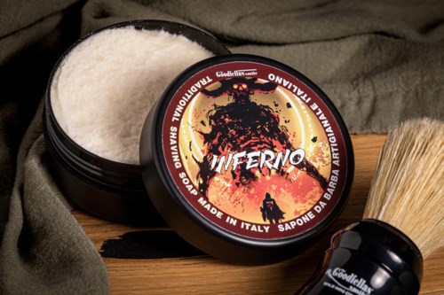 A black container of shaving soap titled 'Inferno' with a fiery illustration. Other grooming items and a cloth are nearby.