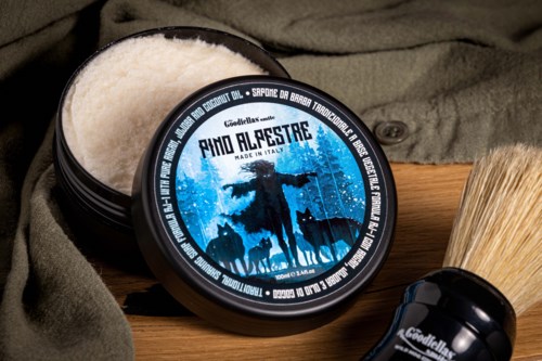 A black jar with blue label, containing shaving cream, and a shaving brush beside it on a cloth surface.