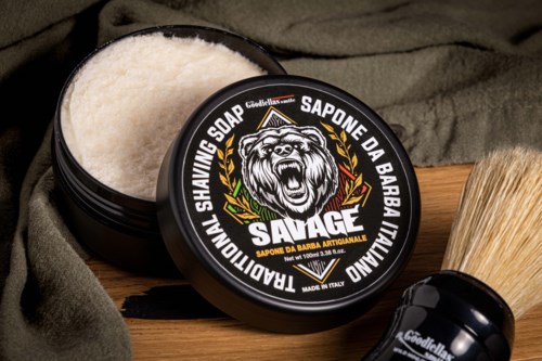 A black container of shaving soap with a lion logo and bold text, partially open, on a wooden surface with a brush.