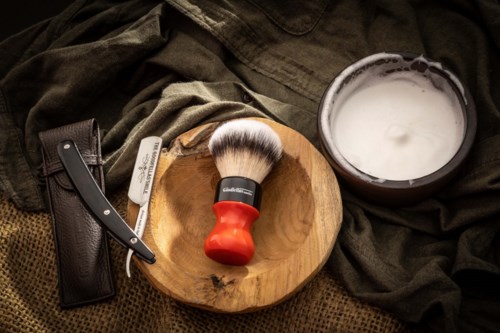 A shaving brush, straight razor, and shaving cream on a wooden plate with dark fabric around.