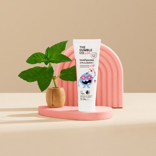 Eine Kinder-Zahnpastatube mit Erdbeergeschmack neben einer kleinen grünen Pflanze in einem Topf, mit einem pinken dekorativen Bogen dahinter.