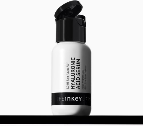 Een witte fles met een zwarte dop, gelabeld Hyaluronic Acid Serum van The Inkey List, op een donkere ondergrond.