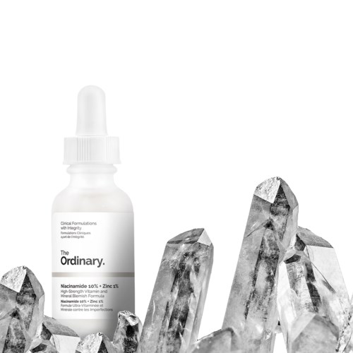 A white bottle with a dropper cap, labeled "The Ordinary," is surrounded by clear quartz crystals.